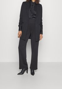 Černý sak s tvarovanými ramenními částmi, kombinovaný s černou košilí s volánkovým detailem a kalhotami s širokými nohavicemi. Černé boty na podpatku.