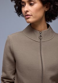 Femme aux cheveux bouclés portant une veste beige texturée avec une fermeture éclair anneau argentée, regardant vers sa gauche sur un fond gris uni.
