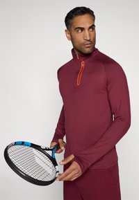 Burgundi pikkade varrukatega sporditopp, millel on neljandikupael ja oranžid aktsendid, koos musta tennisereketiga, millel on sinised detailid.