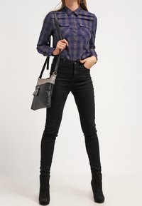 Camicia a quadri blu navy con bottoni, dotata di due tasche sul petto, abbinata a jeans skinny neri e stivaletti; con in mano una piccola borsa nera e grigia.