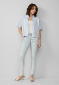 Chemise à manches courtes bleu clair sur un débardeur blanc, associée à un jean skinny en denim clair et à des talons blancs à lanières. Textures lisses et ajustées.