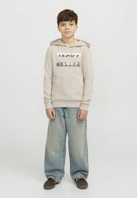 Felpa beige con testo stampato, tasca frontale e cappuccio con cordoncino. Abbinata a jeans blu chiaro a gamba larga e scarpe marroni. Design semplice.