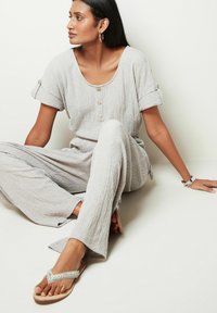 Jumpsuit grigio chiaro texturizzato con maniche corte e dettagli in bottone, abbinato a ciabatte decorate. Design semplice e rilassato.