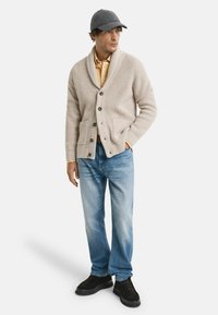 Man med grå keps, beige kofta med sjalkrage och knappar, ljusbrun skjorta, blå jeans och svarta skor, stående med en hand i fickan.