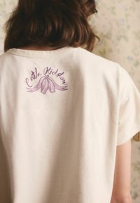 Młoda osoba z brązowymi włosami do ramion nosi kremową koszulę z fioletowym napisem "Cath Kidston" i wzorem kokardki na plecach, w pobliżu szyi.