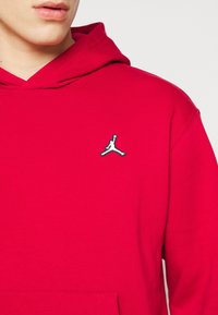 Personne portant un sweat à capuche rouge vif avec un logo Jumpman noir et blanc sur la poitrine, visible du menton jusqu'en haut du torse.