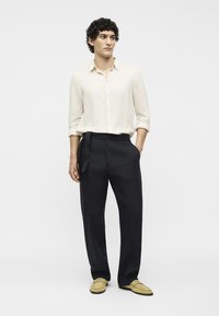 Άνδρας με κατσαρά μαλλιά που φοράει μια μπεζ πουκαμίσα με κουμπιά, μαύρο παντελόνι με κορδόνι στο πλάι και ανοιχτό καφέ μοκασίνια, στέκεται μπροστά από μονόχρωμο φόντο.