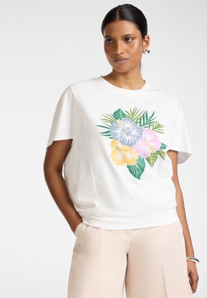 Donna che indossa una camicia bianca a maniche corte con stampa floreale multicolore, pantaloni beige, braccialetto d'argento e orecchini a cerchio, posando con la mano in tasca.