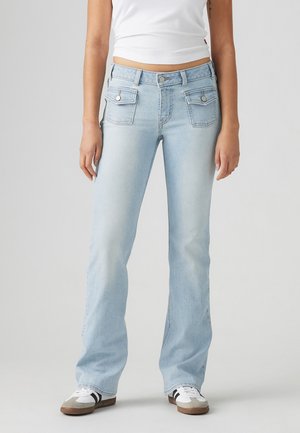 Kobieta w jasnoniebieskich jeansach typu bootcut z przednimi kieszeniami z klapkami, białym topie i białych butach sportowych z czarnymi paskami.