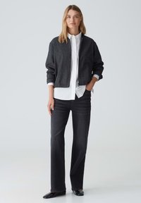 Grau bomberjacke mit gerippten Bündchen über einem weißen Hemd mit Kragen, kombiniert mit geraden schwarzen Jeans und schwarzen Slippern. Glatte Texturen.