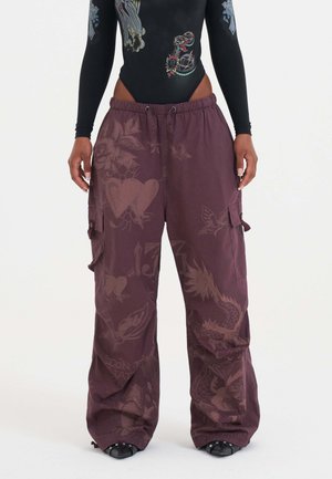 Vrouw in paarse wijde cargobroek met tattoo-geïnspireerde prints en een zwarte langekous bodysuit met kleurrijke ontwerpen.