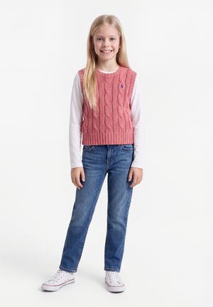 Junges Mädchen mit langen blonden Haaren, das eine rosa Zopfmusterweste, ein weißes Langarmshirt, blaue Jeans und weiße Turnschuhe trägt, steht und lächelt.