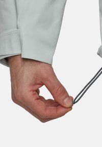 Manche de veste gris clair avec un détail de poignet. Une main tient un cordon de serrage gris foncé pour un ajustement, mettant en valeur un élément de conception fonctionnel.