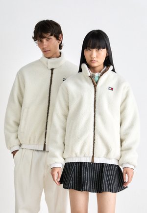 Cream-colored fleece jackets with high collars, front zippers, and ribbed cuffs. One features an embroidered logo; the other is paired with a patterned skirt.