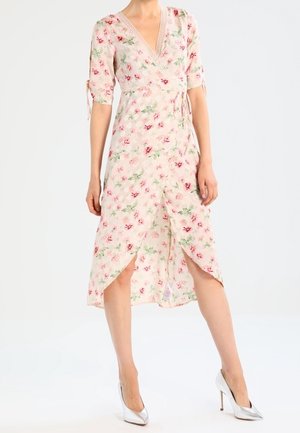 Floral midikleid aus hellrosa Stoff mit kurzen Ärmeln, V-Ausschnitt, vorderen Schlitzen und einem gebundenen Detail in der Taille. Kombiniert mit silbernen Schuhen.