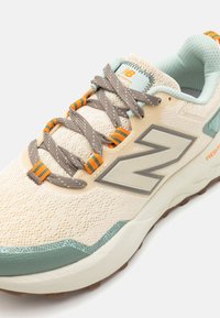 Beige atletiske sneakers med mesh-overdel, mintgrønne accenter og tekstureret gummisål. Har grå snørebånd med orange og grønne striber.