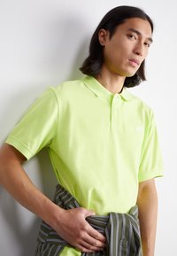 Jasne limetkovozelené polo tričko z bavlny. Obsahuje špicatý golier a krátke rukávy. Na ľavej strane hrude je biele logo Nike. Okolo pása je viazaná vertikálne pruhovaná látka.