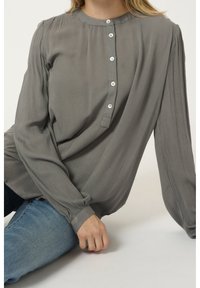 Femme portant un chemisier gris à manches longues avec des boutons blancs, associé à un jean bleu, assise devant un fond uni.