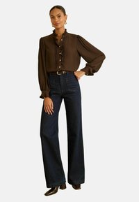 Donna che indossa una blusa marrone con rouches e bottoni a forma di cuore, jeans scuri a gamba larga, cintura marrone e scarpe con tacco, in piedi con una mano sui fianchi.