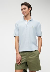 Polo à manches courtes bleu clair en tissu texturé, avec un col à trois boutons et un petit logo vert en accent. Associé à un short vert olive.