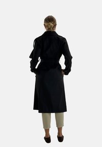 Czarny płaszcz trench z paskiem w talii, długimi rękawami i bocznymi rozcięciami, wykonany z gładkiego materiału, noszony na jasne spodnie.