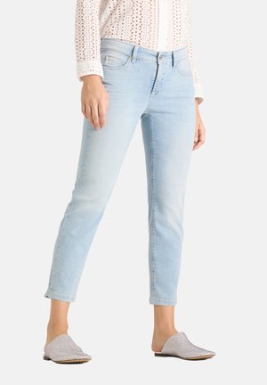 Frau trägt hellblaue Slim-Fit-Jeans, weißes langärmeliges Blusentop mit Lochmuster und glitzernde Slipper, steht mit entspannten Armen.