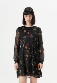Robe noire avec de longues manches transparentes à pois, un corsage ajusté et une jupe évasée, ornée d'un motif floral en rouge, jaune et vert.