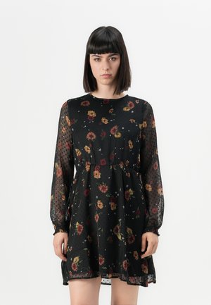 Robe noire avec de longues manches transparentes à pois, un corsage ajusté et une jupe évasée, ornée d'un motif floral en rouge, jaune et vert.