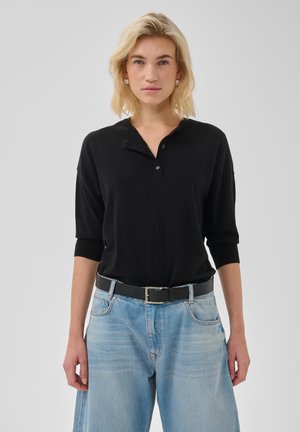 Person med skulderlangt blondt hår har sort knap-bluse på, blå jeans med sort bælte og står med fronten mod kameraet foran en ensfarvet lys baggrund.