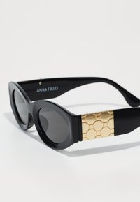 Black sunglasses with rounded lenses, a glossy finish, and gold hexagonal-patterned accents on the temples. Brand name embossed on the side.