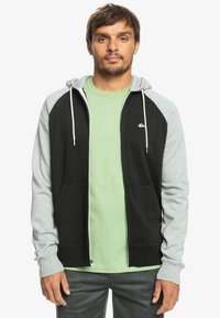 Čierna a svetlo šedá zapínacia mikina s priliehavým strihom, nastaviteľnou kapucňou so šnúrkou a dvoma prednými vreckami, cez svetlo zelené tričko.