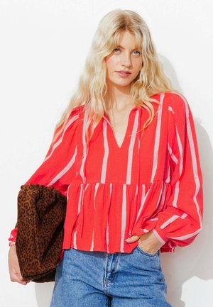 Femme blonde portant un chemisier rayé rouge et blanc à manches longues et un jean bleu, tenant une pochette marron à imprimé léopard.