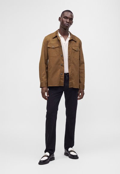Homme portant une veste marron boutonnée sur une chemise blanche, un pantalon noir et des mocassins noir et blanc, debout devant un fond uni.