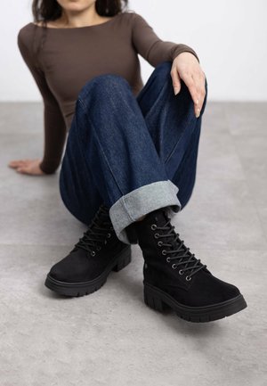 Botas de ante negras hasta el tobillo con suelas gruesas y un notable detalle de cordones, combinadas con jeans de mezclilla oscura remangados y una blusa marrón ajustada.
