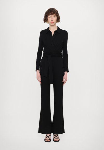 Jumpsuit noir ajusté avec manches longues, fermeture à boutons à l'avant, ceinture enroulée à la taille, jambes évasées et texture lisse. Associé à des sandales noires à lanières.