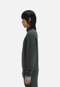 Sudadera de color verde oscuro con cuello alto, con un acento azul en los puños. Textura suave y corte relajado. Se muestra vista lateral.