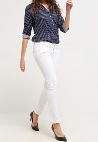 Kvinna som går, klädd i en marinblå blus med prickar, vita slim-fit byxor och svarta högklackade skor mot en ljus, enkel bakgrund.