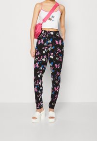 Pantalones joggers negros con estampado de flores en rosa, azul y blanco, combinados con un top corto blanco y un bolso rosa. La modelo lleva sandalias de plataforma blancas.