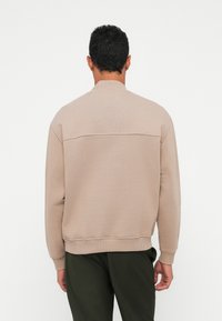 Beige mikina s texturovaným povrchem, žebrovaným límcem a manžetami, s vodorovným švem přes zadní stranu. Na sobě má tmavě zelené kalhoty.