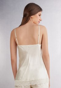 Satinbaby - camisol i elfenben med tunna justerbara band och spetsdetalj vid fållen, med en slät textur och en åtsittande silhuett.