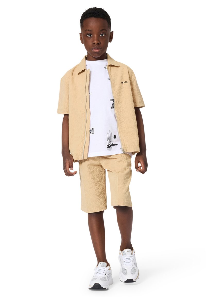 Dreng iført beige kortærmet jakke og matchende shorts, hvid grafisk t-shirt og hvide sneakers stående mod en hvid baggrund.