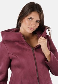 Giacca zip-up borgogna con colletto alto, caratterizzata da una texture di tessuto liscia e un design elegante. La cerniera e le tasche sono anch'esse visibili.