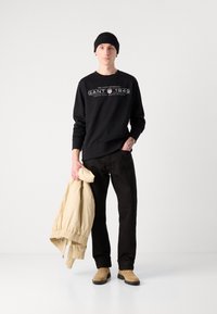 Svart hettegenser med hvit logo, svarte bukser, beige dunjakke og brune ankelstøvler. Modellen har på seg en svart strikket lue. Enkel bakgrunn.