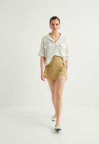 Vit stickad polo-liknande topp med korta ärmar och två framfickor, kombinerad med khaki shorts med hög midja och sidfickor.
