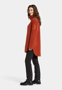 Orange fleece pullover med høje krave og sideslids, der har en struktureret finish og et logo-patch på venstre ærme. Bæres med sorte jeans.