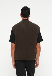 Άνδρας φορώντας ένα χήρο καφέ αμάνικο πλεκτό γιλέκο πάνω από ένα μαύρο κοντομάνικο μπλουζάκι, στέκεται να κοιτάει από πίσω σε λευκό φόντο.