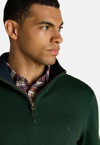 Zelený pulóver so zipsom a golierom, s námorníckym akcentom a modrým logom. Pod ním je vidieť károvanú košeľu v červených, bielych a modrých farbách.