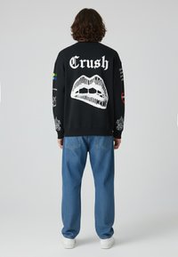 Felpa nera con il testo bianco "Crush" e una grafica di labbra sul retro. Maniche lunghe con vari stampati. Abbinata a jeans azzurri chiari.
