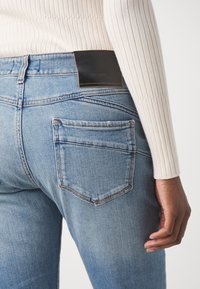 Svetle modré džíny z denimu s klasickým designem zadní kapsy, černou koženou nášivkou na pase a žebrovaným krémovým svetrem.