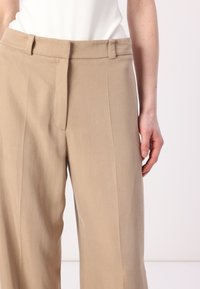 Beige op maat gemaakte broeken met een voornaad en riemlussen, gemaakt van lichte stof, met een strak ontwerp en minimalistische zakdetails.